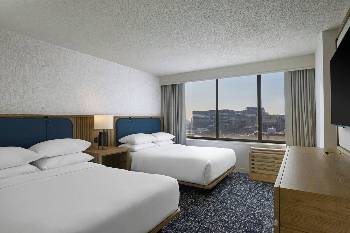Habitación de hotel con dos camas, un escritorio, televisión y un ventanal con vistas a la ciudad.