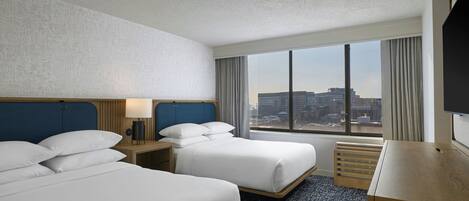 A hotel room with two beds, a desk, a TV, and a large window with a city view.