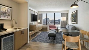 A modern hotel room with a kitchenette, a dining area, a seating area with a sofa and chairs, a flat-screen TV, and large windows offering a city view. - Sheraton Philadelphia University City Hotel (Philadelphia)