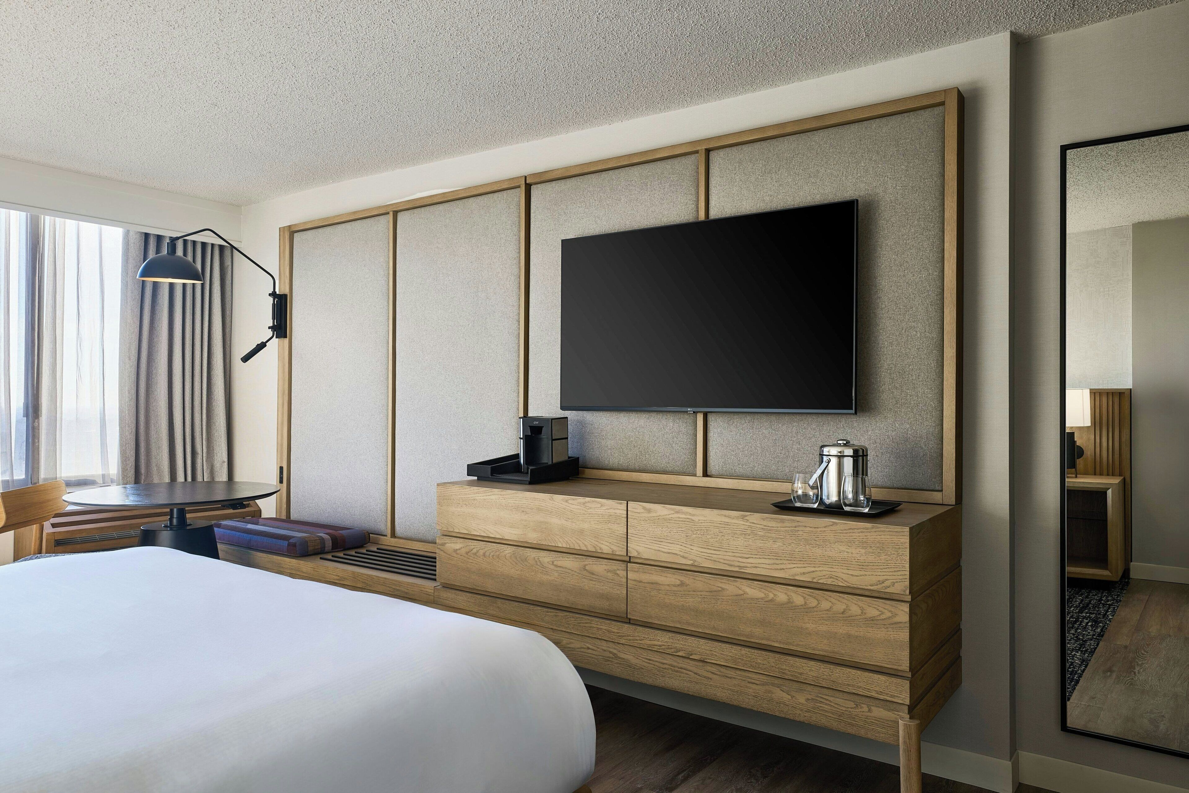 A hotel room with a bed, a flat-screen TV mounted on a wooden panel, a coffee maker, and a desk with a lamp.