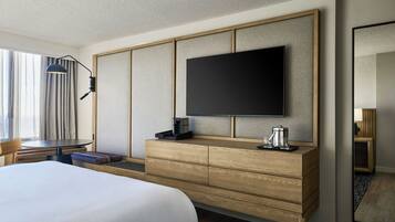 A hotel room with a bed, a flat-screen TV mounted on a wooden panel, a coffee maker, and a desk with a lamp.