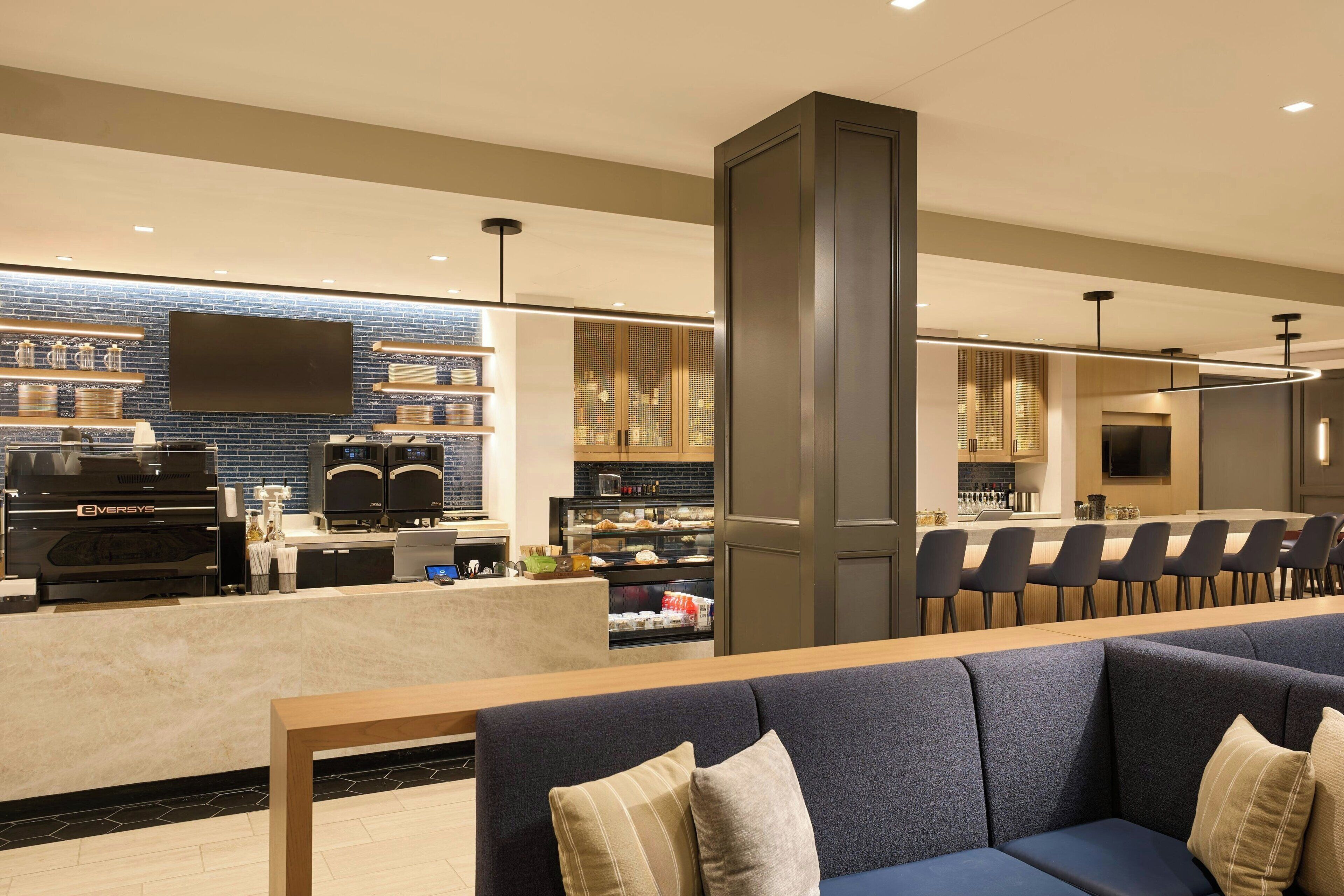 A modern hotel lobby with a seating area, a bar, and a kitchen in the background.