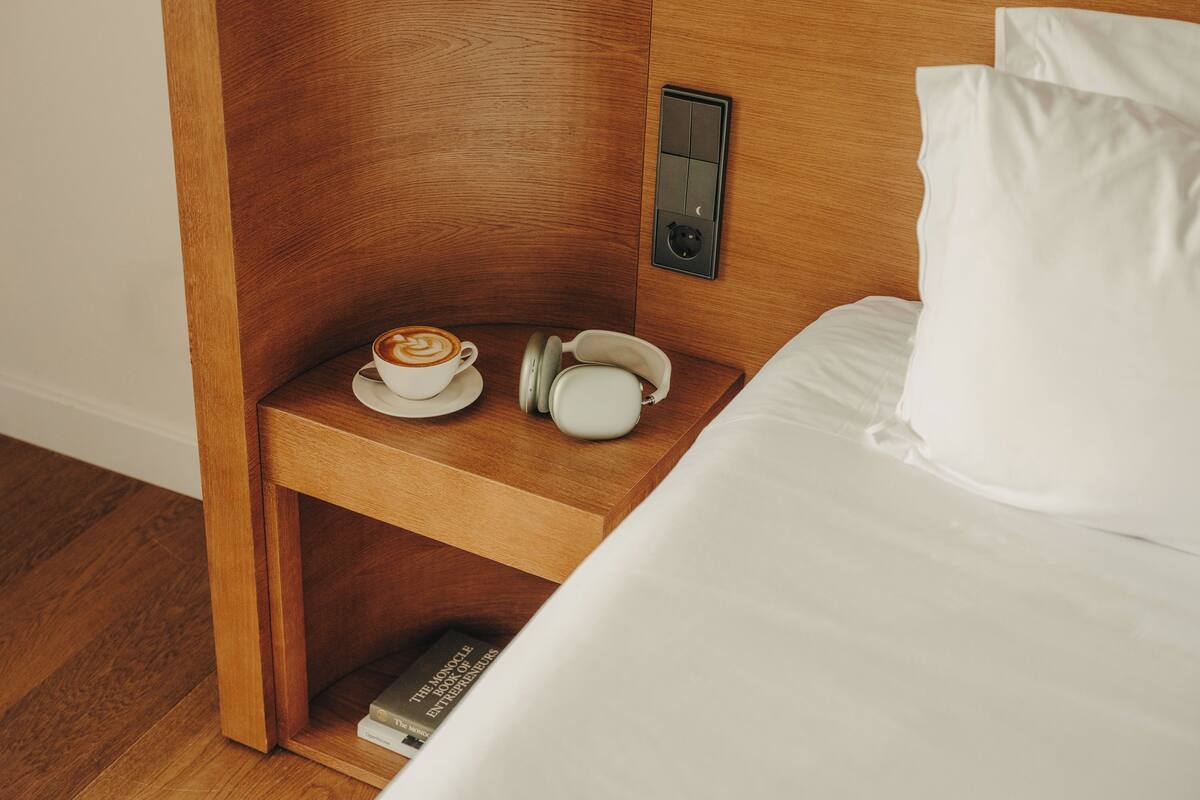 A wooden bedside table with a cup of coffee, a white pillow, and a book titled 'The Monocle Book of Entrepreneurs.'