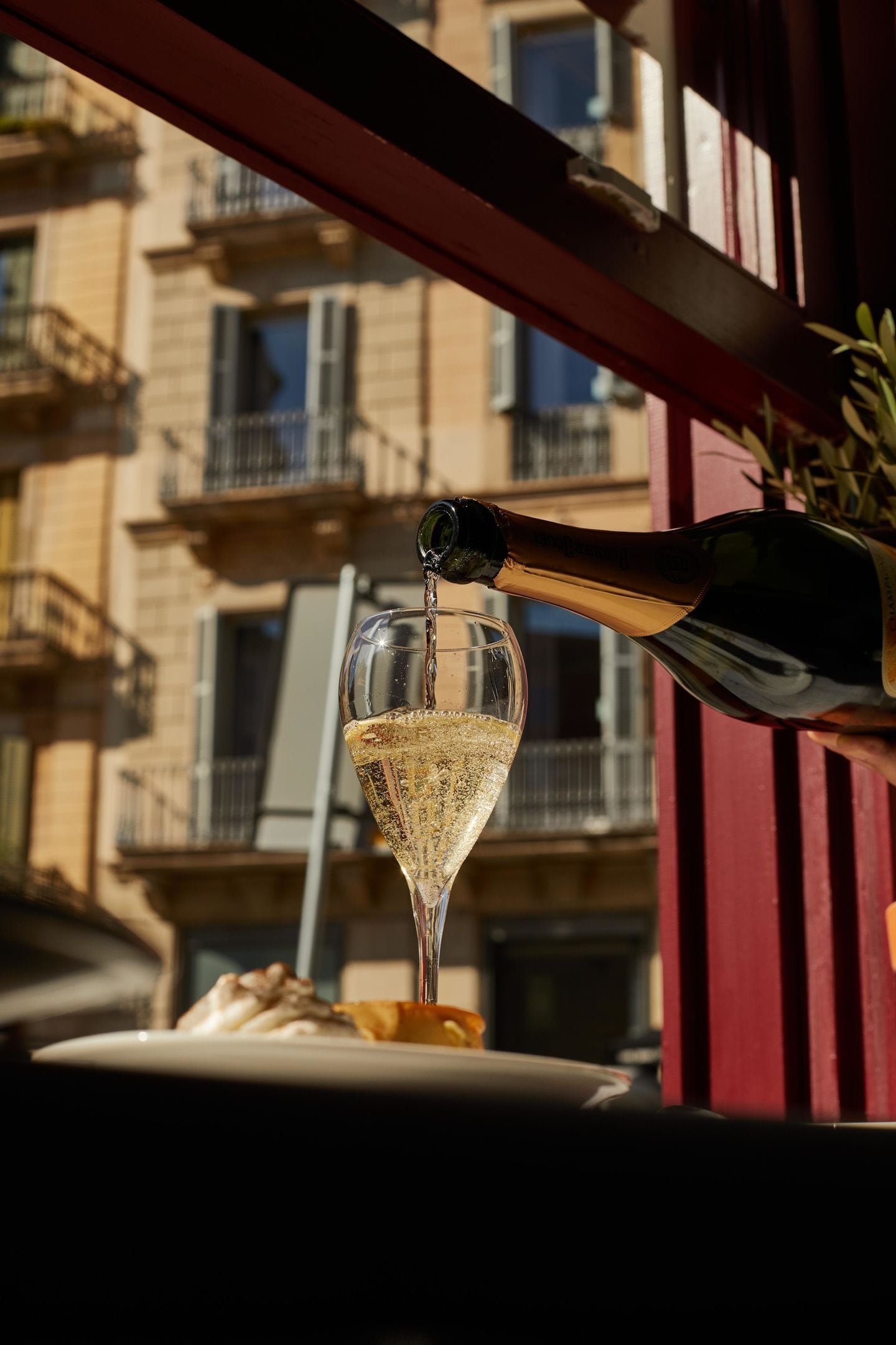 A balcony with a glass of champagne being poured, a plate with food, and a building with balconies in the background.