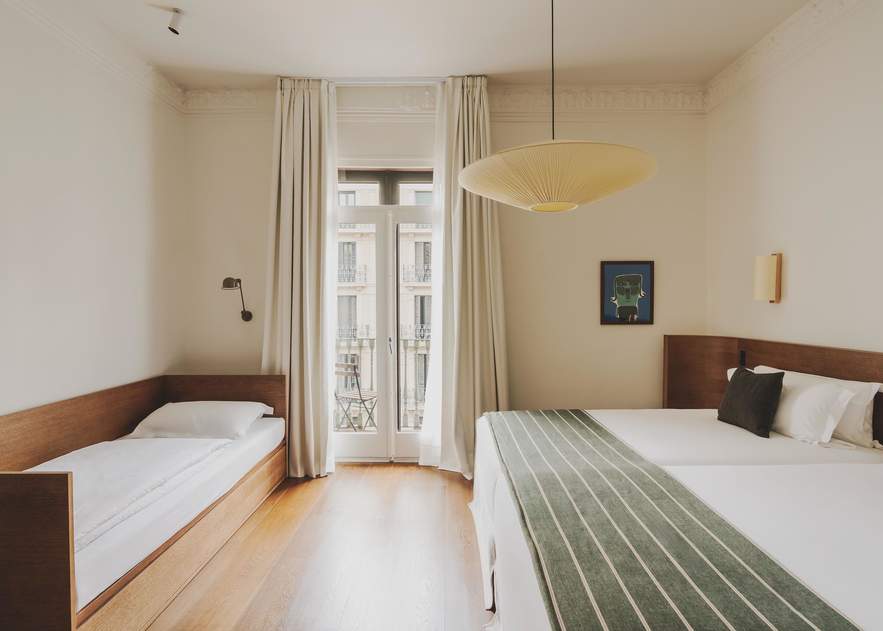 A hotel room with two single beds, a large window with curtains, a hanging light fixture, and a framed picture on the wall.