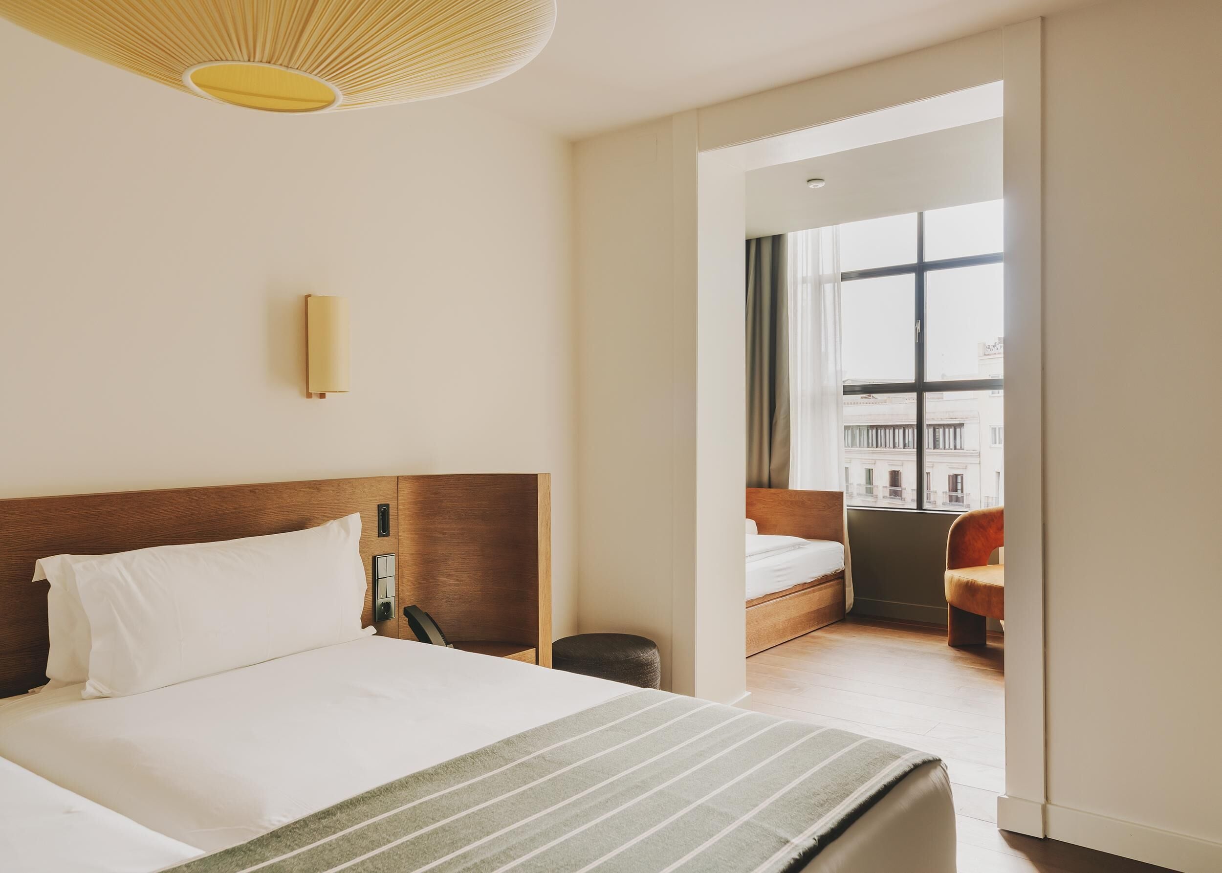 A hotel room with a large bed, a wooden headboard, a bedside table, a lamp, and a view of the city through a window.