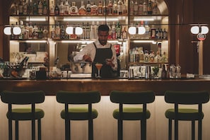 Uma área de bar com um bartender preparando uma bebida, prateleiras com diversos tipos de garrafas e banquetas verdes.