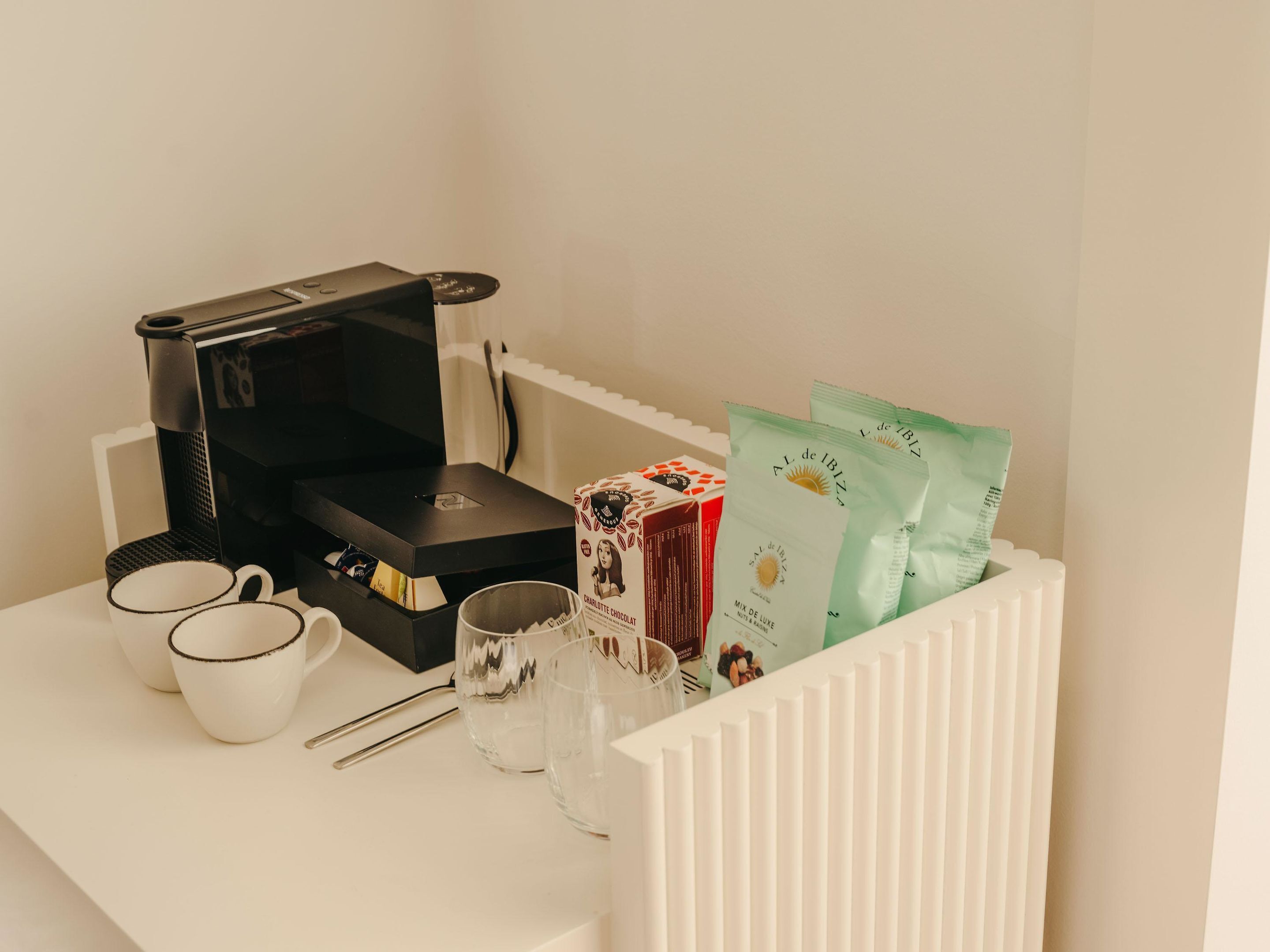 Eine Kaffeemaschine, zwei Tassen, ein Glas, eine Schokoladenschachtel und zwei Teepäckchen auf einem weißen Tisch.