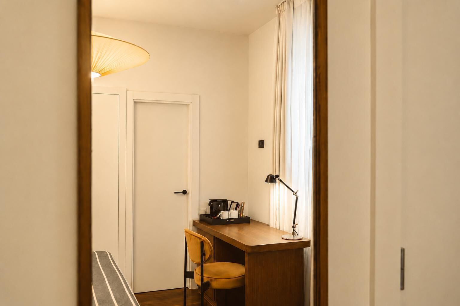 A narrow hallway with a door, a desk with a chair, a lamp, and a window with curtains.