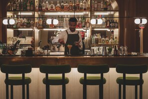 A bar area with a bartender preparing a drink, shelves with various bottles, and green bar stools. - Hotel Regina Barcelona (Barcelona)