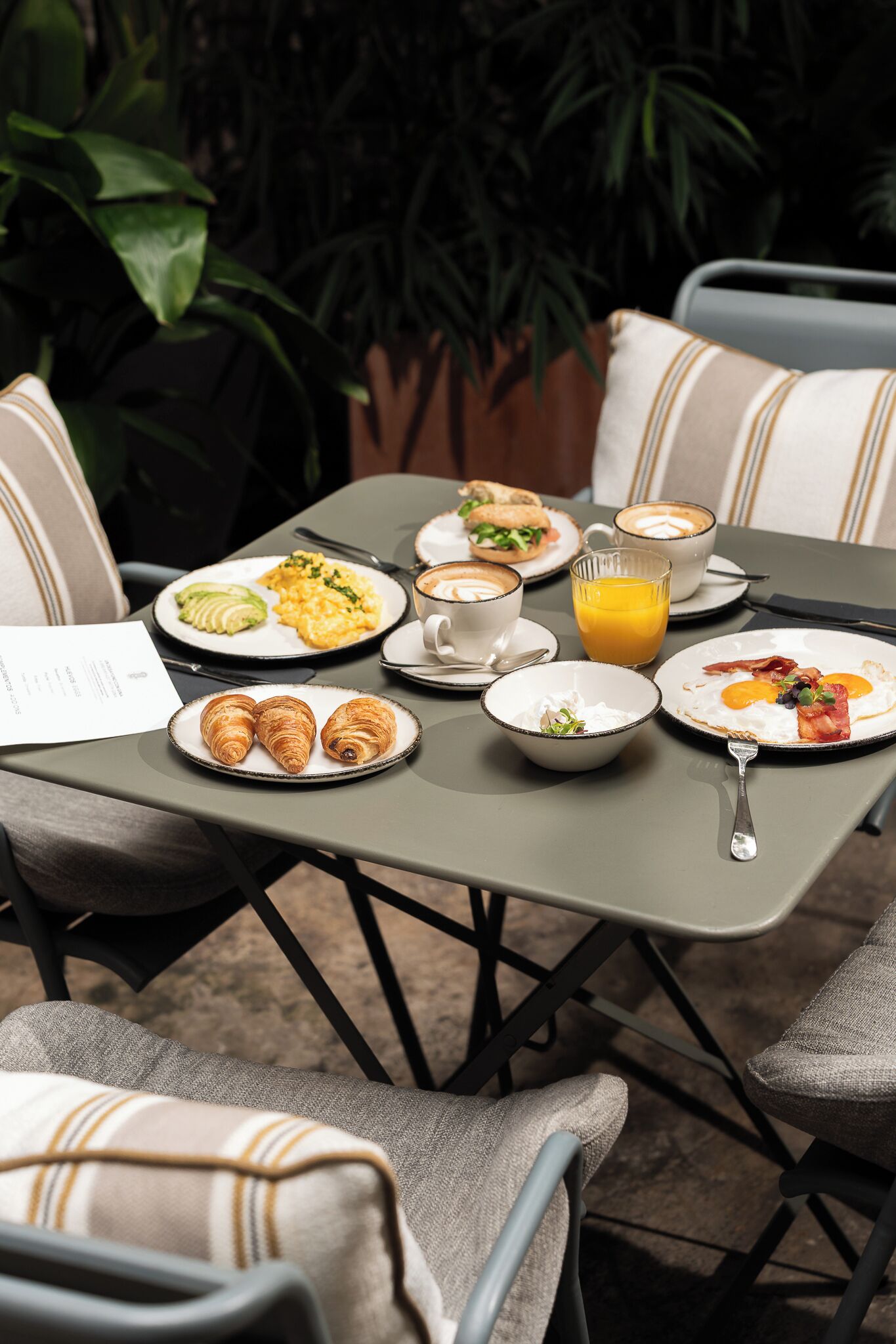 Una mesa dispuesta con desayuno, incluyendo cruasanes, huevos, tocino y zumo de naranja, rodeada de sillas y una planta en maceta.