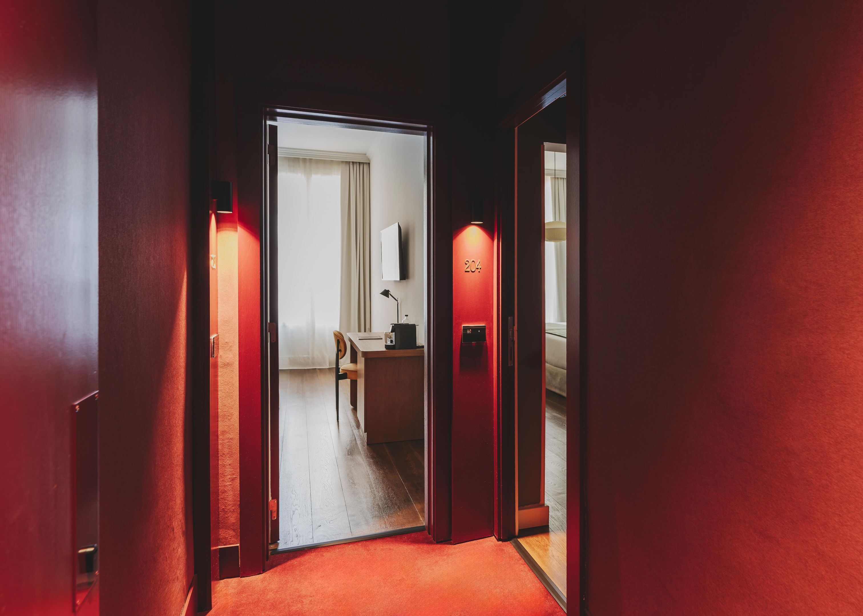 A hotel hallway with a red carpet, a desk with a lamp, and a room number 204 visible through an open door.