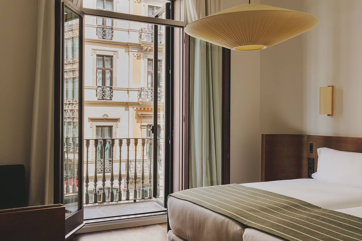 A hotel room with a large window, a bed, a bedside table, and a hanging light fixture.