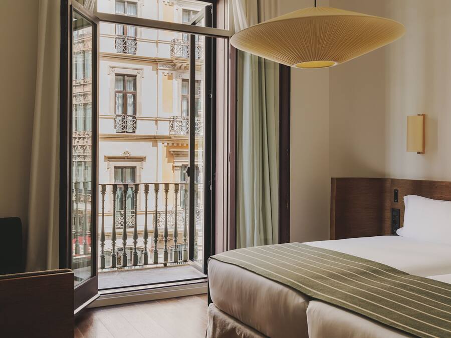 A hotel room with a large window, a bed, a bedside table, and a hanging light fixture.