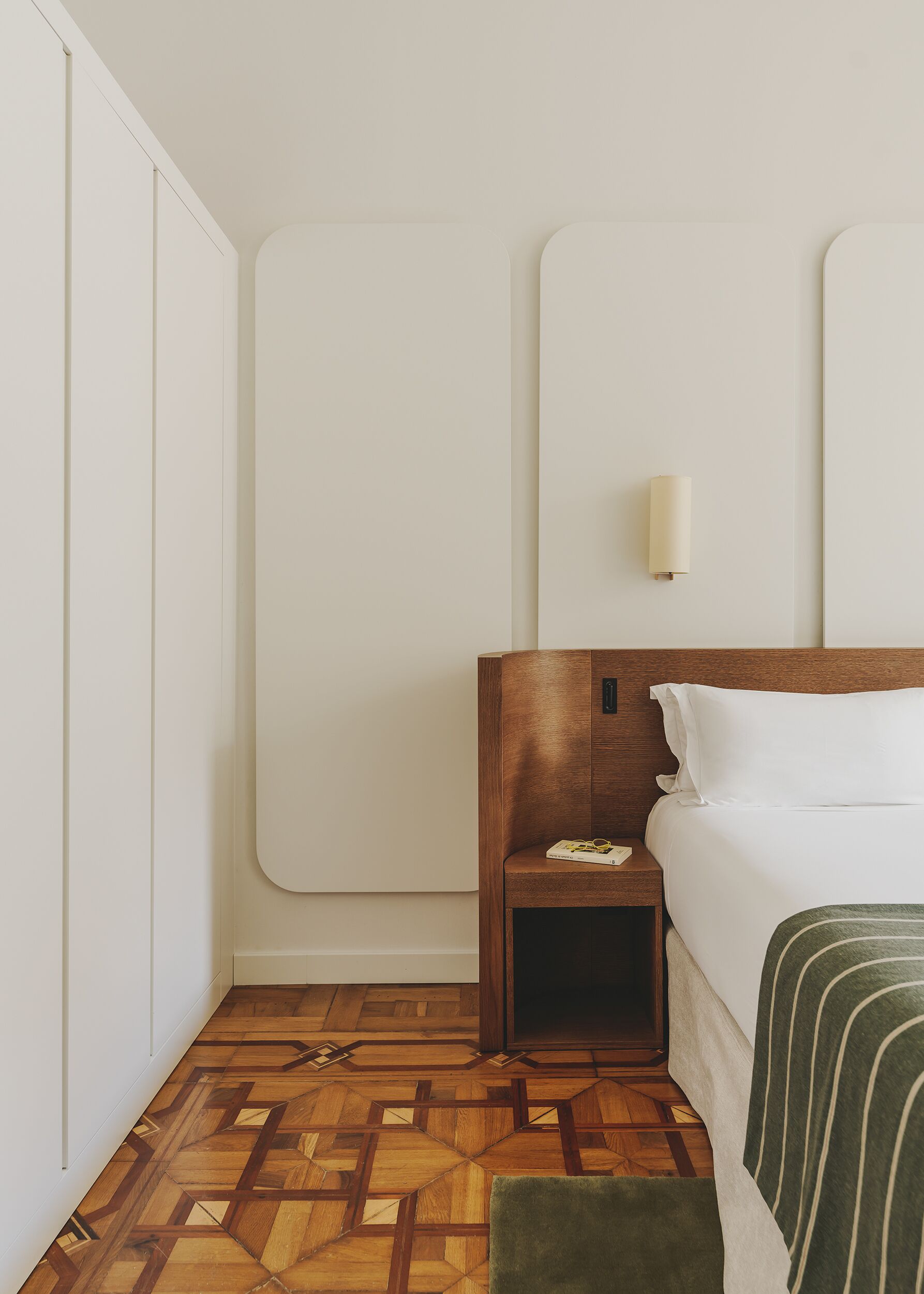 A hotel room with a wooden headboard, a bedside table with a book and glasses, a green throw pillow, and a patterned wooden floor.