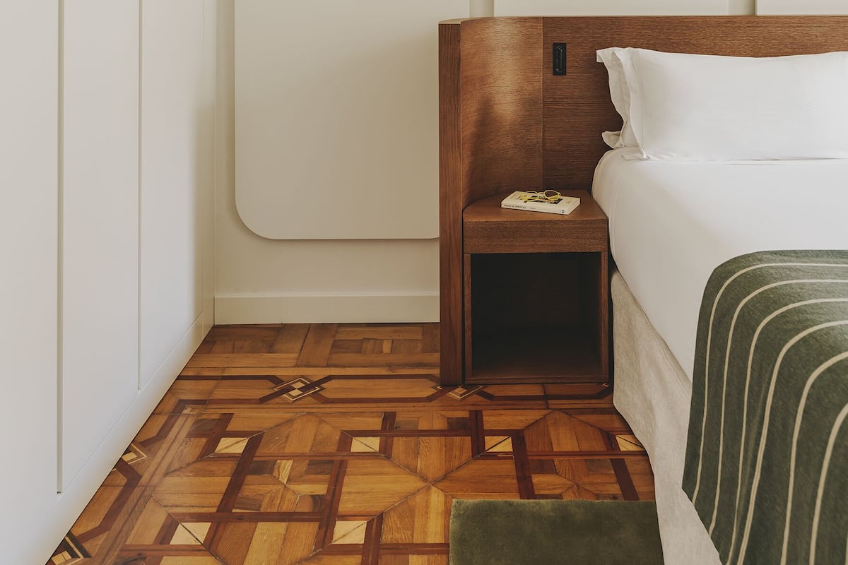A hotel room with a wooden headboard, a bedside table with a book and glasses, a green throw pillow, and a patterned wooden floor.