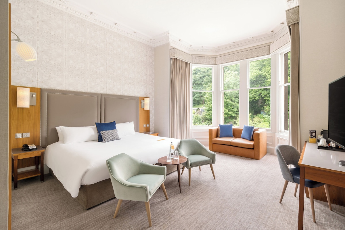 A hotel room with a large bed, a sitting area with a sofa and armchairs, a desk with a television, and a window with a view of greenery.