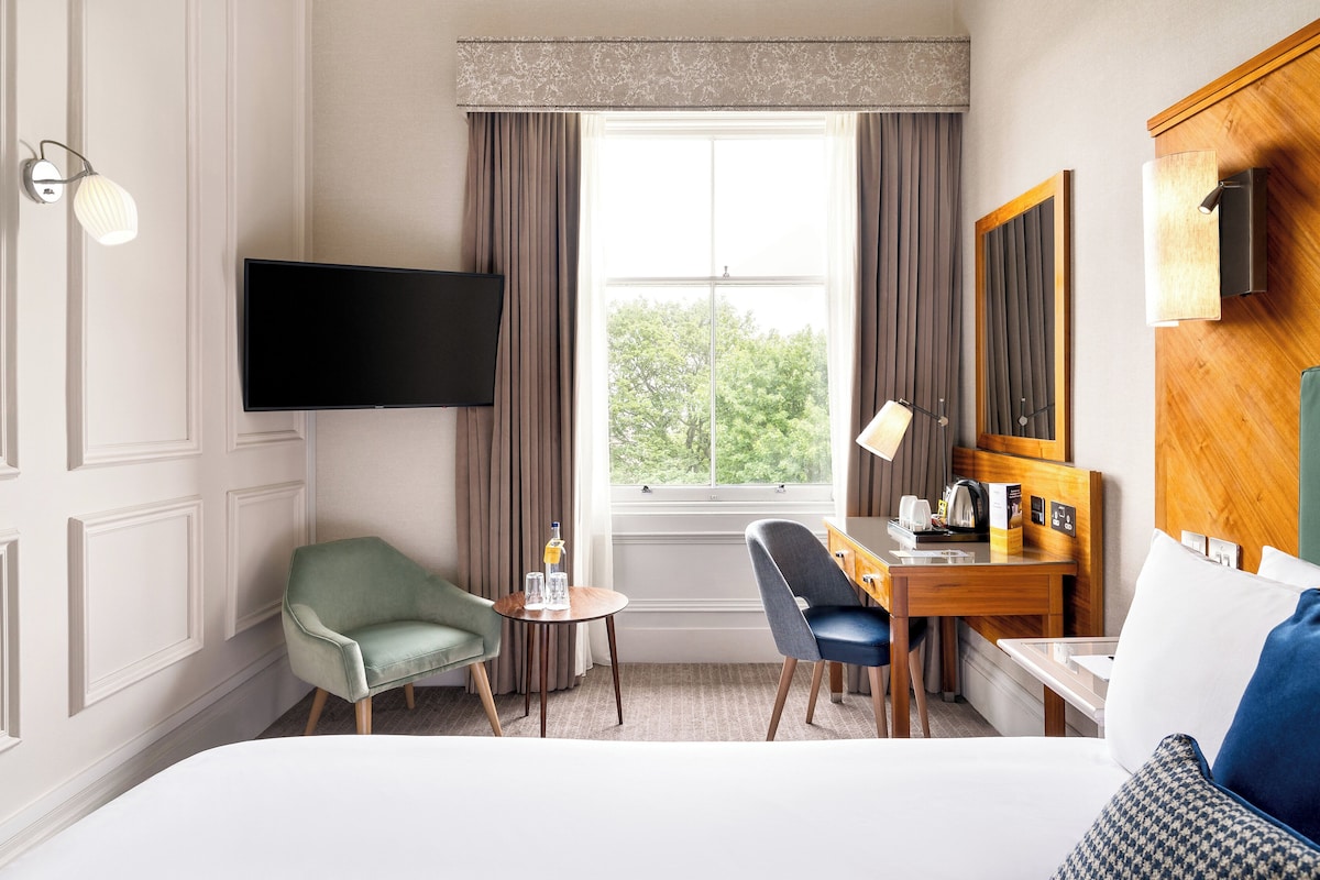 A hotel room with a bed, two chairs, a small table with a bottle and glasses, a desk with a coffee maker, a flat-screen TV, and a window with curtains.