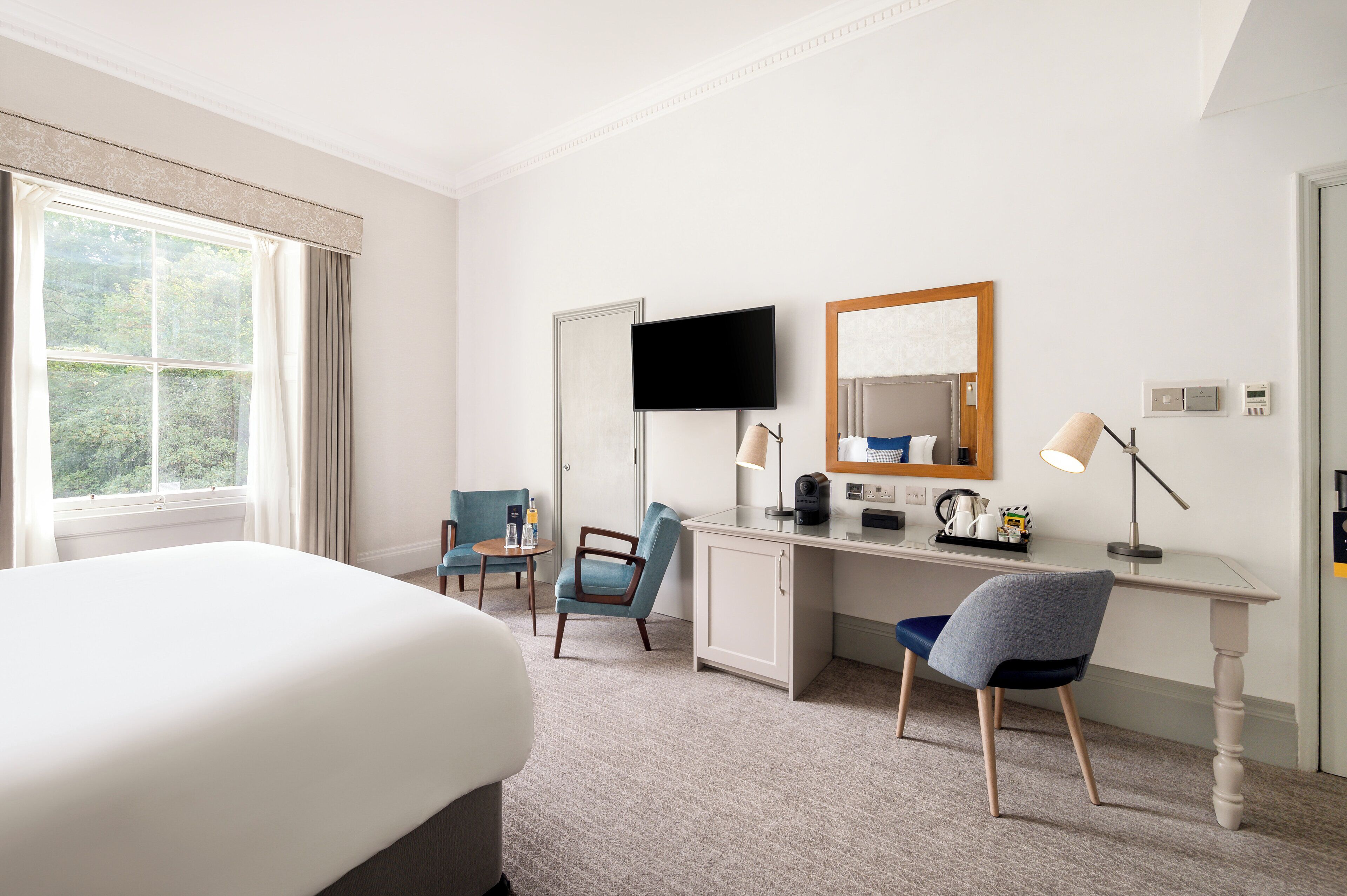 A hotel room with a bed, a desk with a computer, a chair, a TV, and a window with curtains.
