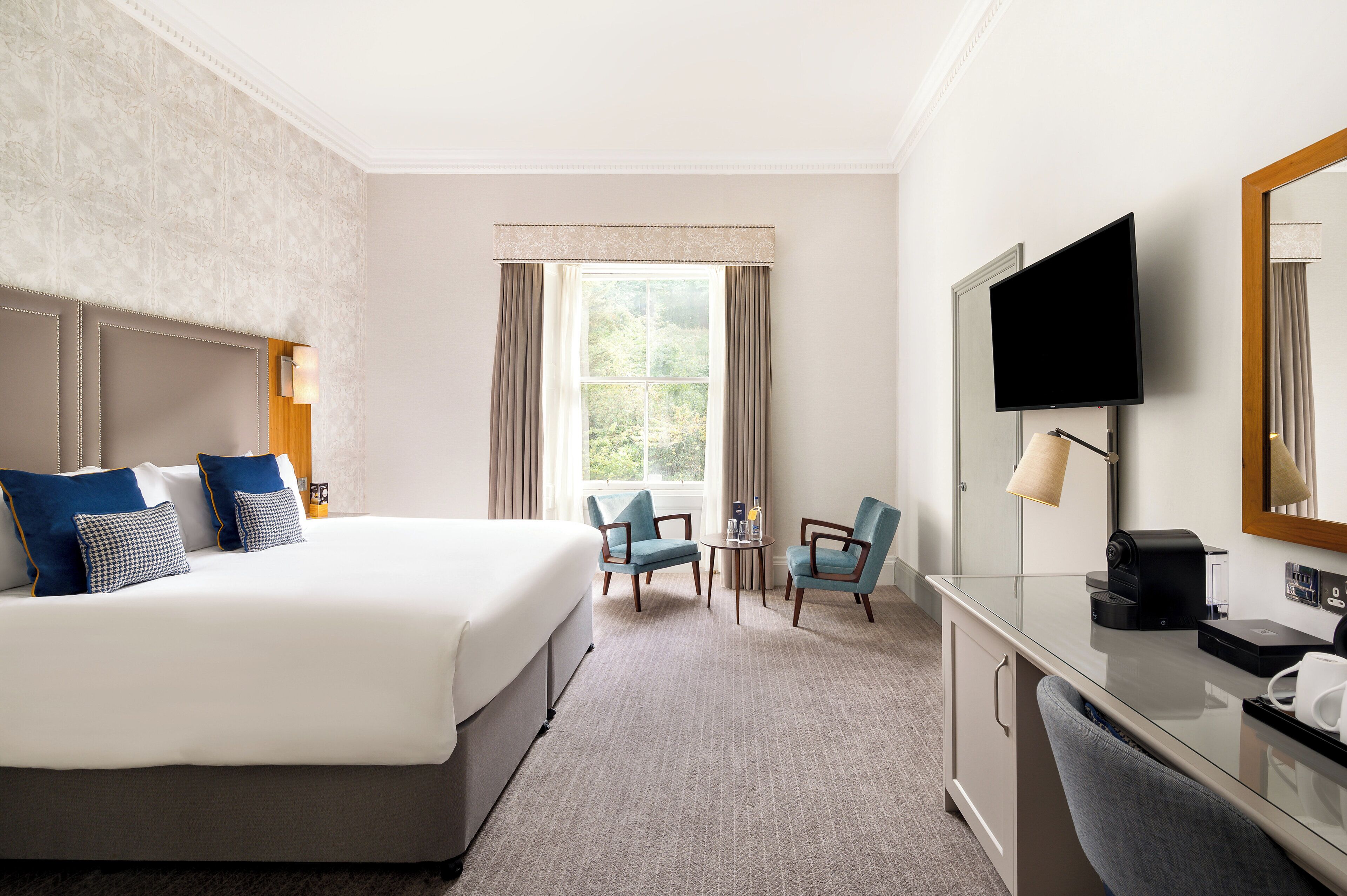 A hotel room with a large bed, two armchairs, a small table, a TV, and a desk with a mirror.