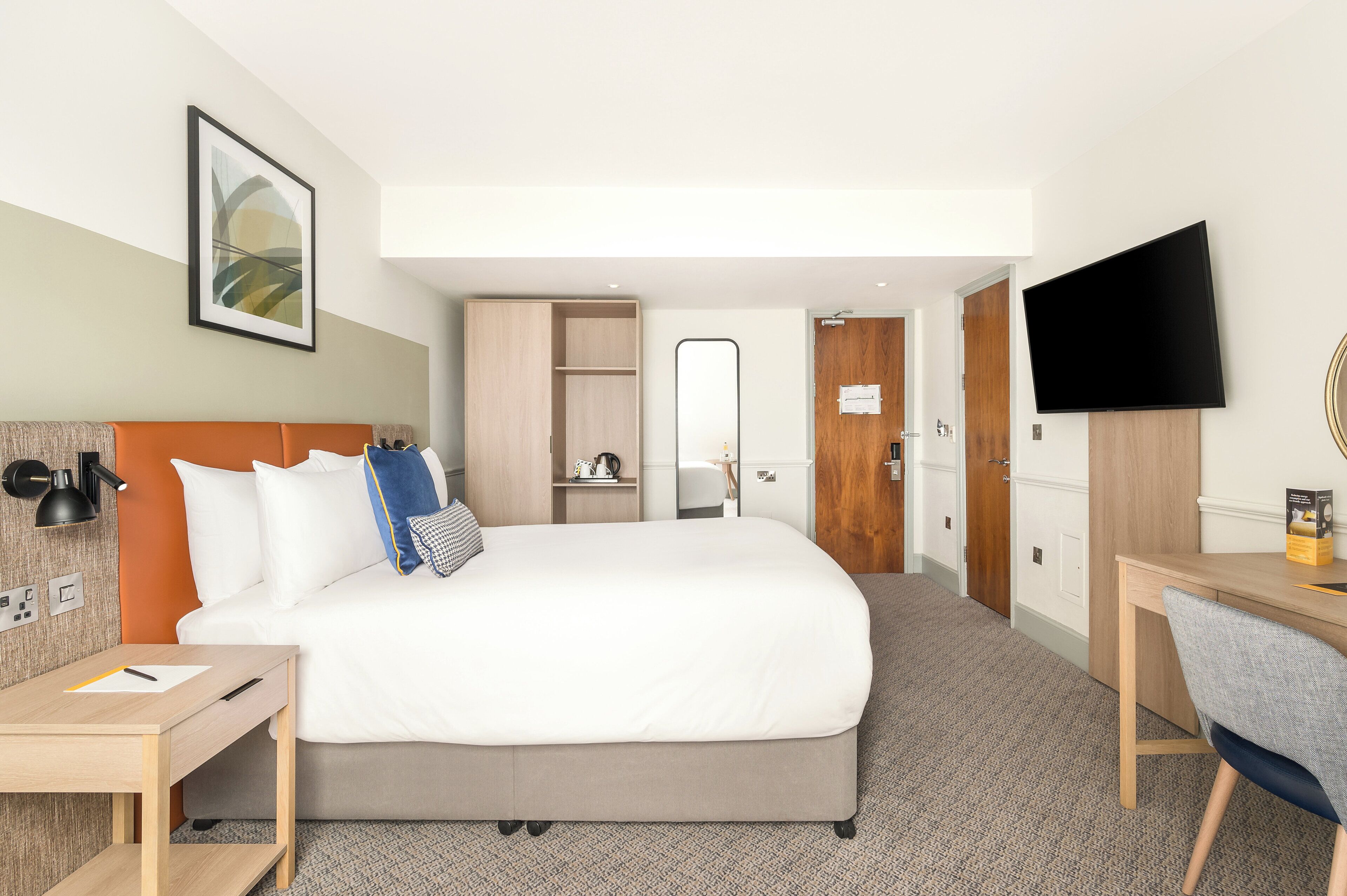 A hotel room with a bed, desk, chair, TV, wardrobe, and a mirror.