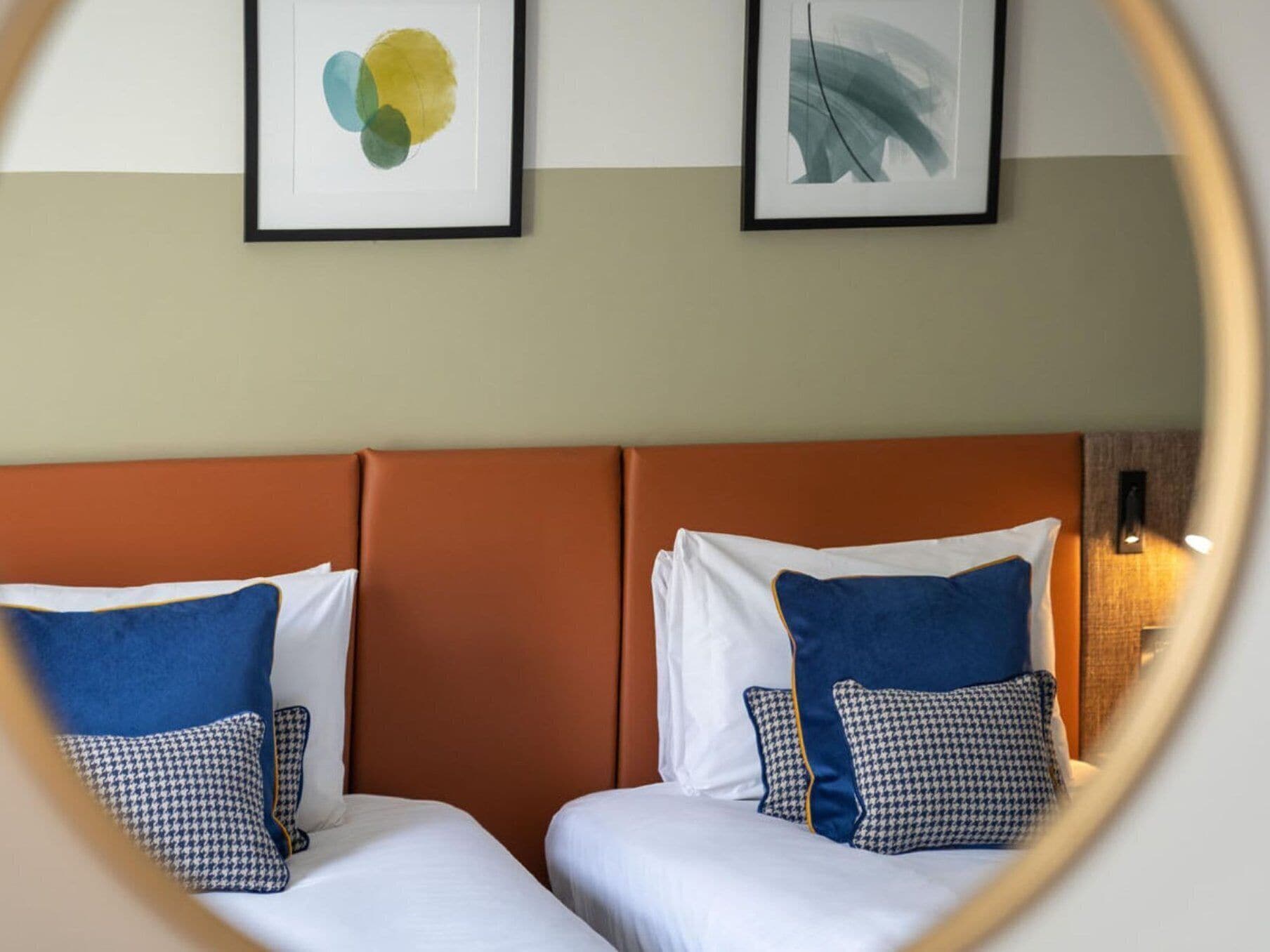 A hotel room with two beds, each with white bedding and blue pillows. There are abstract paintings on the wall above the beds.