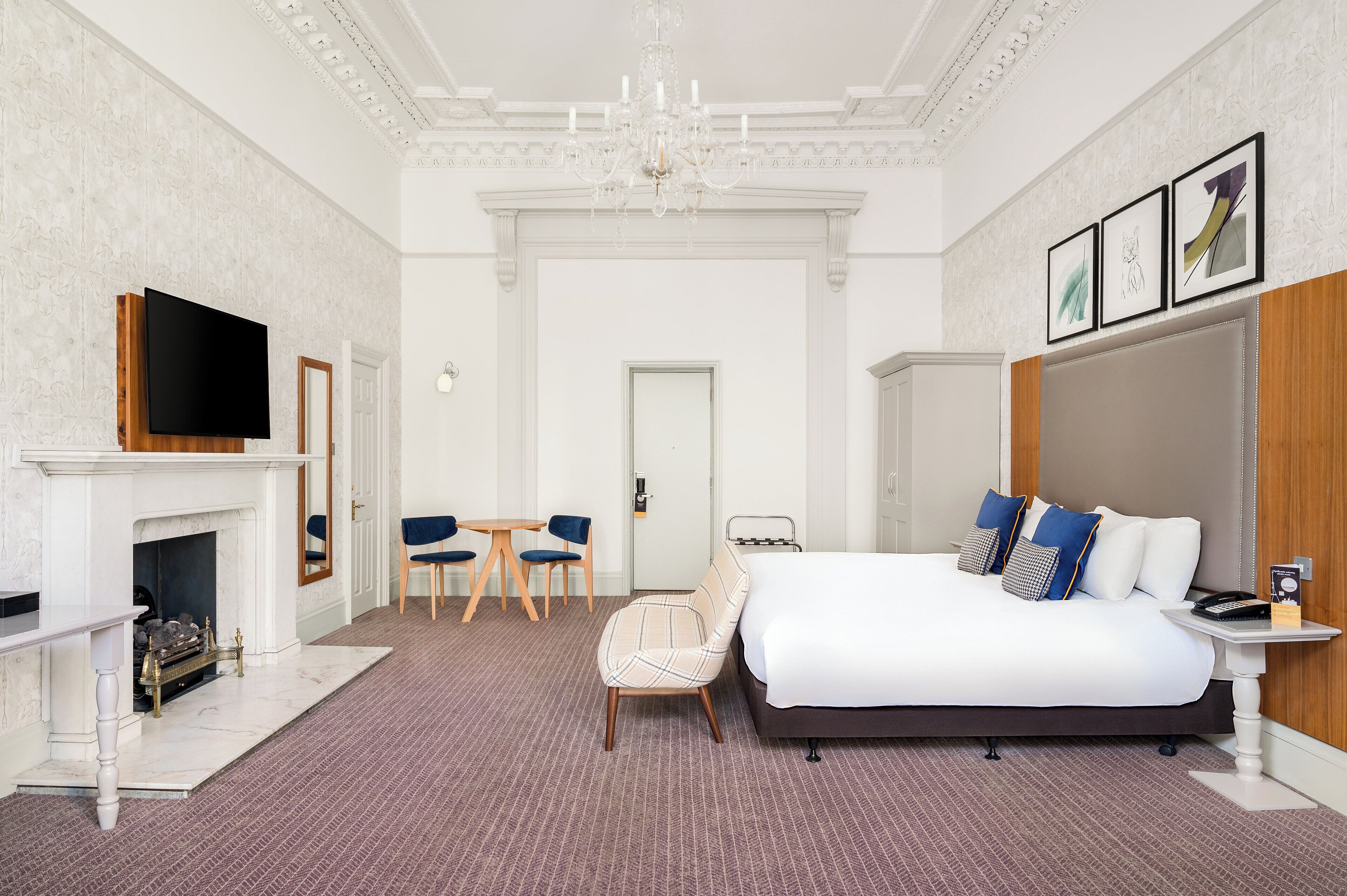 A hotel room with a large bed, a TV, a fireplace, a dining area, and a chandelier.