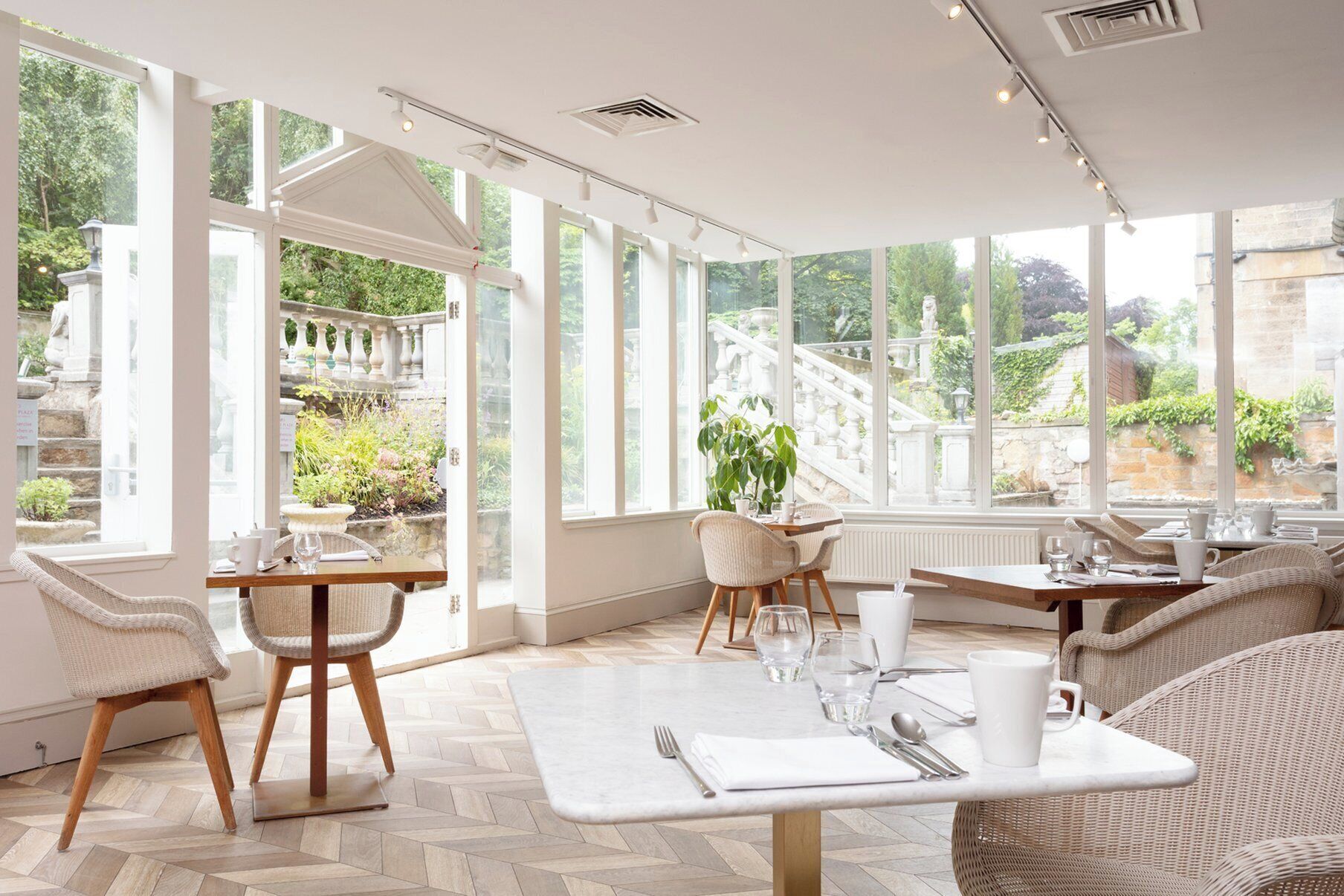 A bright, sunlit room with large windows, wicker furniture, and a herringbone floor pattern.