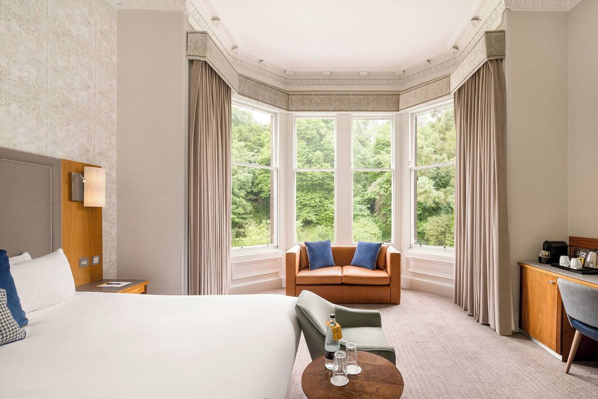 A hotel room with a large bed, a sitting area with a sofa and armchairs, a desk with a chair, and a view of greenery through large windows.