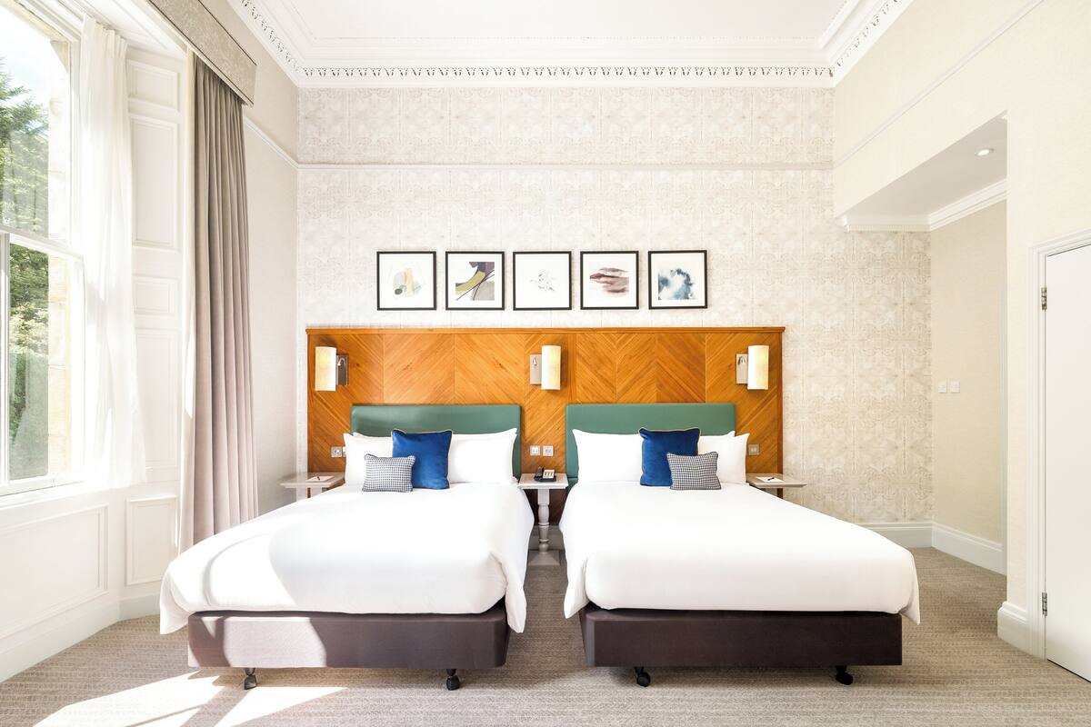 A hotel room with two beds, a wooden headboard, a window with curtains, and a wall with framed pictures.
