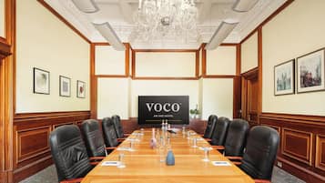 A conference room with a long wooden table, black leather chairs, a chandelier, and a large screen displaying "VOCO AN IHG HOTEL".