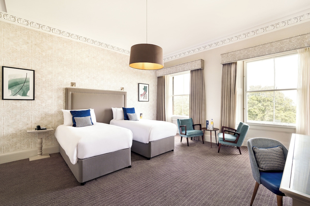 A hotel room with two beds, a sitting area with a table and chairs, and large windows.