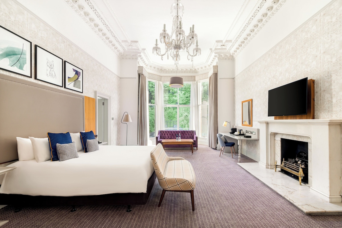 A spacious hotel room with a large bed, a seating area, a fireplace, and a television.