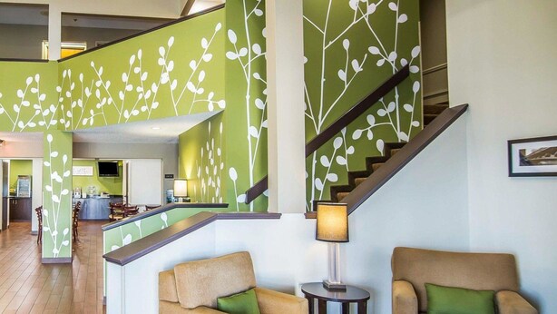 A lobby with a staircase, two beige armchairs with green cushions, a small table with a lamp, and a reception desk.