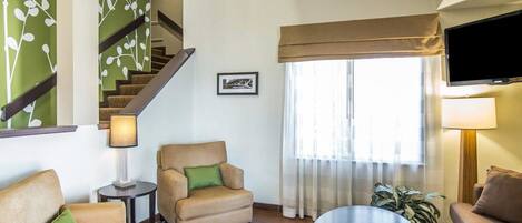 A living room with a sofa, armchair, and a coffee table. There is a staircase with a green wall and a framed picture on the wall.