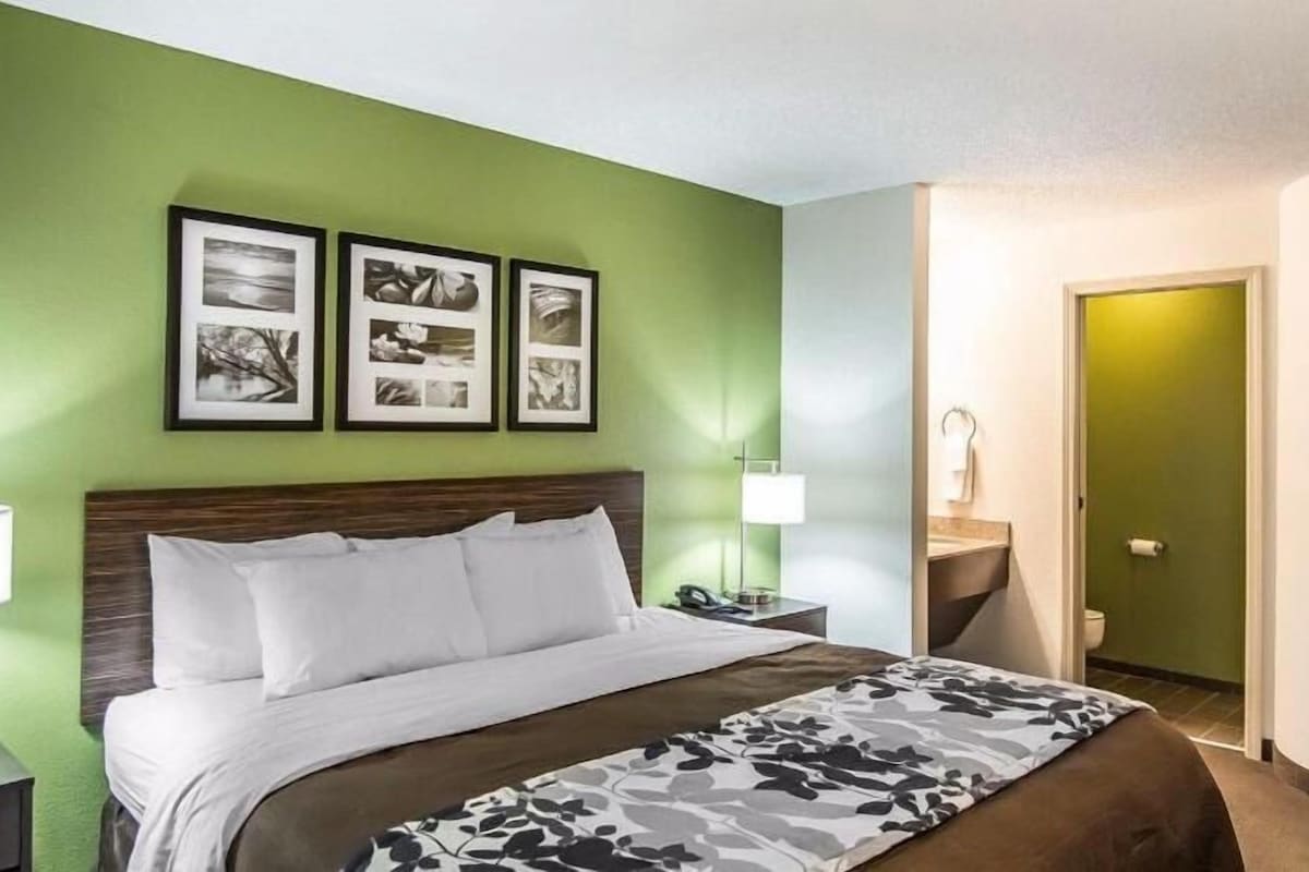 A hotel room with a bed, green walls, framed pictures, a nightstand, a lamp, and a bathroom visible through an open door.