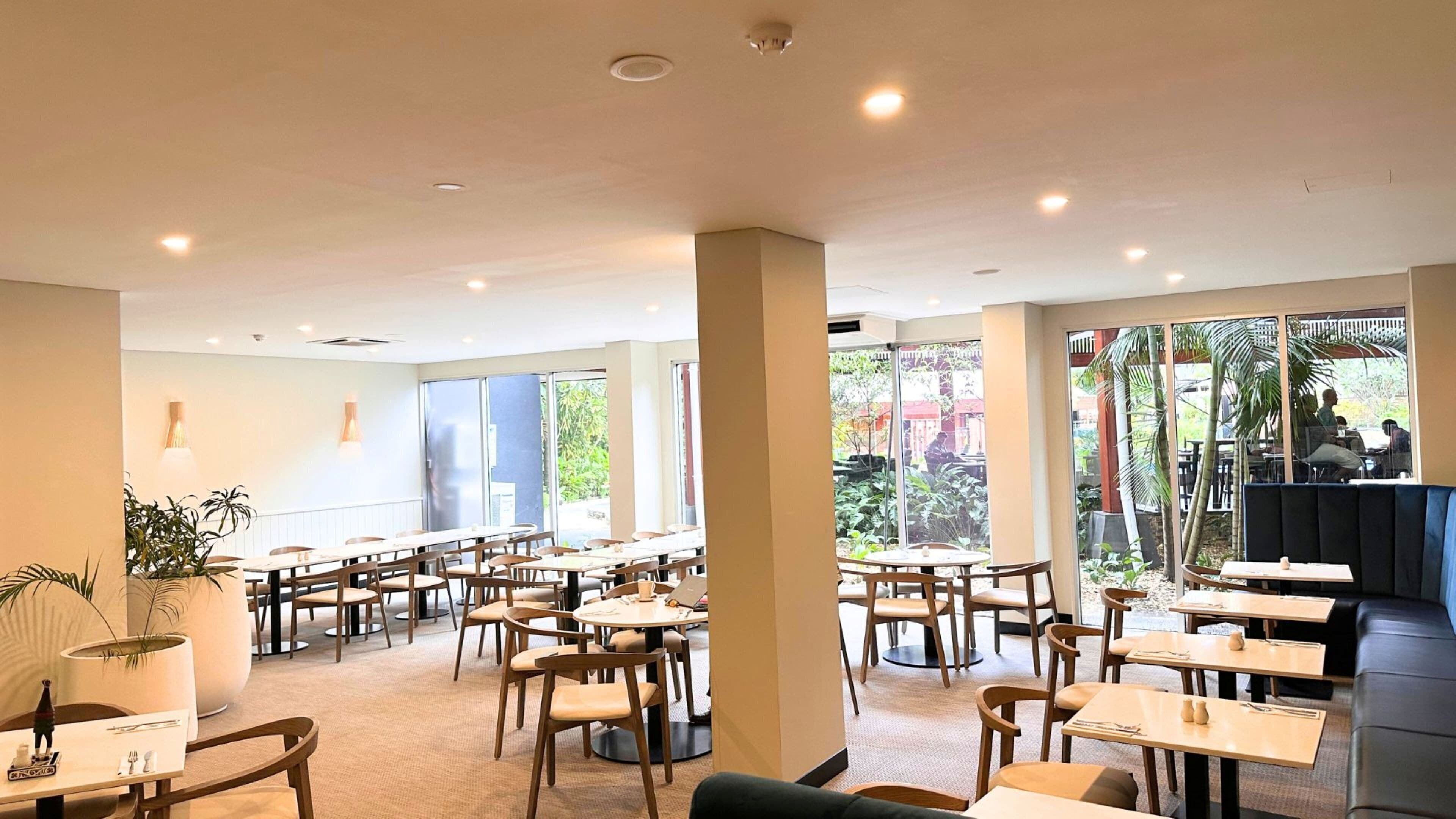 A spacious indoor dining area with multiple tables and chairs, large windows offering views of the outdoors, and a modern ceiling design.