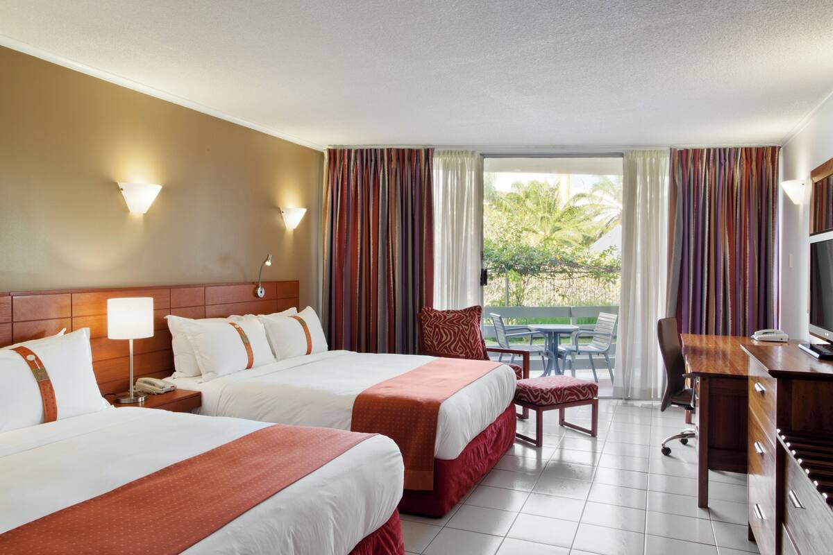 A hotel room with two beds, a desk, a chair, and a balcony with curtains.