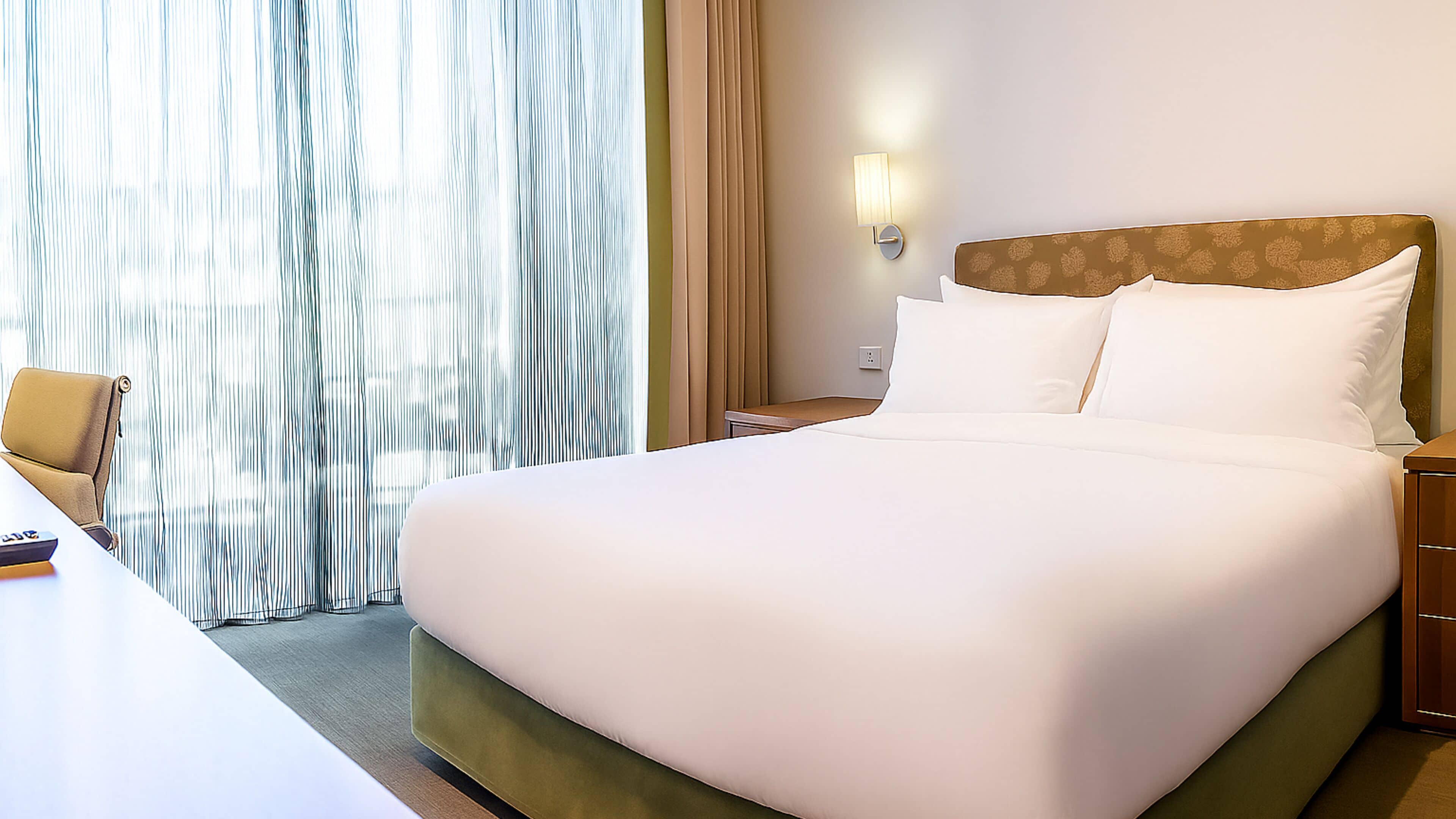 A hotel room with a large bed, a bedside table, a chair, a desk, and a window with curtains.