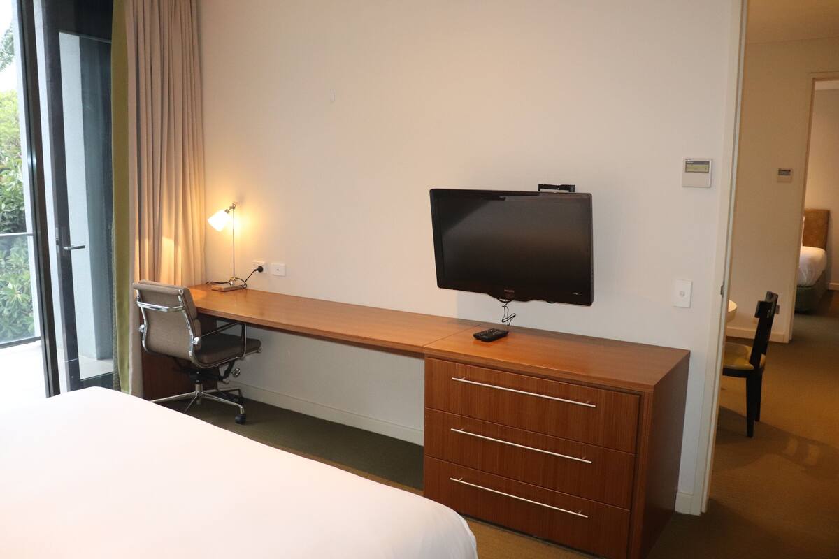 A hotel room with a bed, a desk with a chair, a TV mounted on the wall, and a window with curtains.