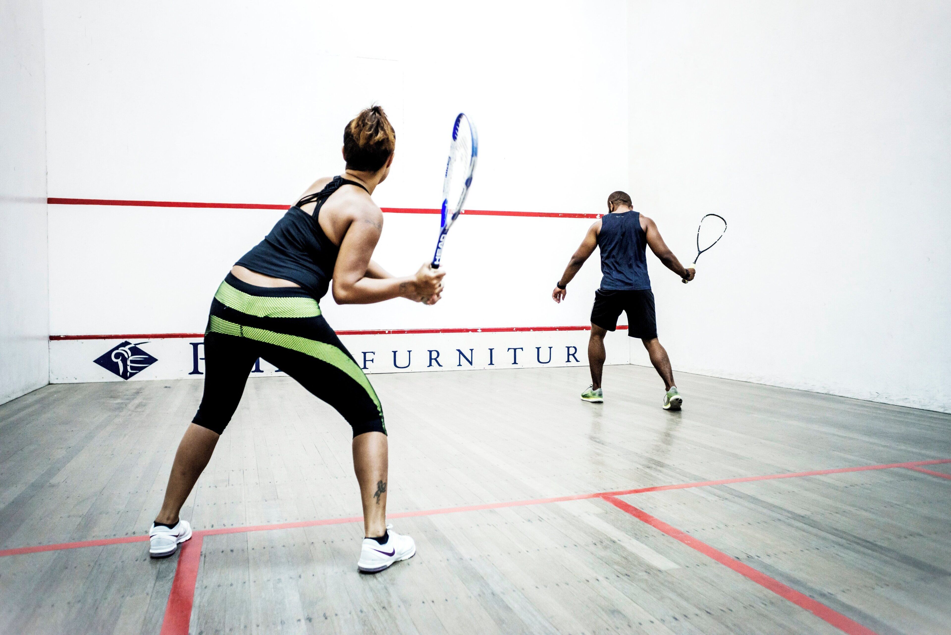 To personer spiller squash i et rum med trægulv og en hvid væg med røde linjer.