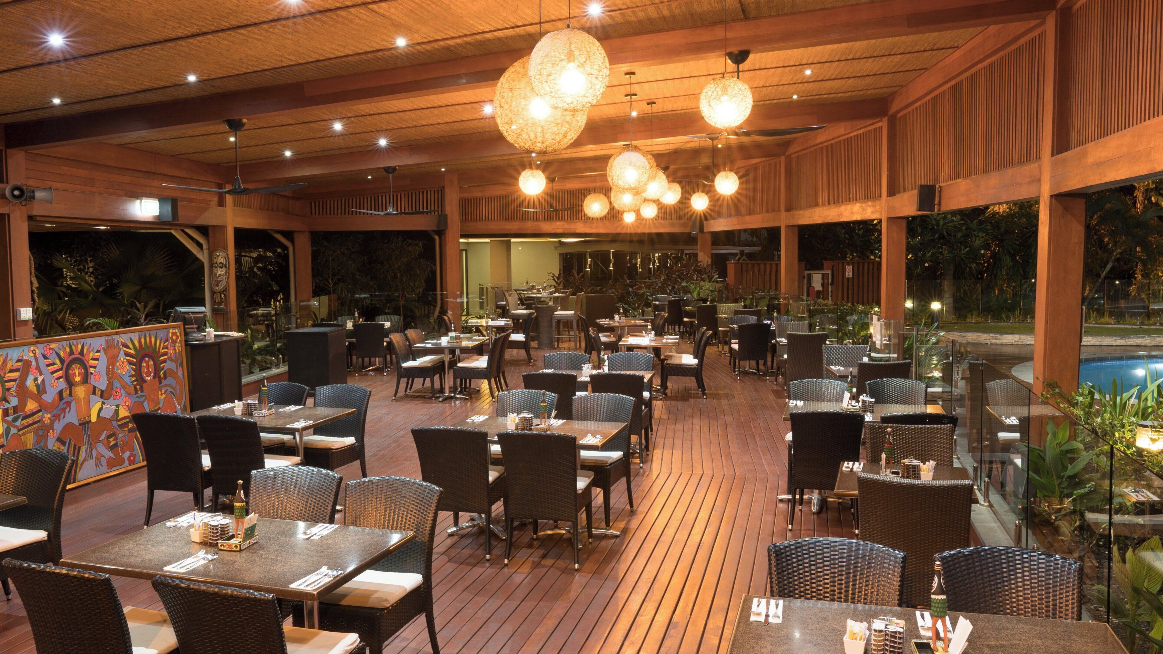 An outdoor restaurant with multiple tables and chairs, wooden flooring, and hanging spherical light fixtures.