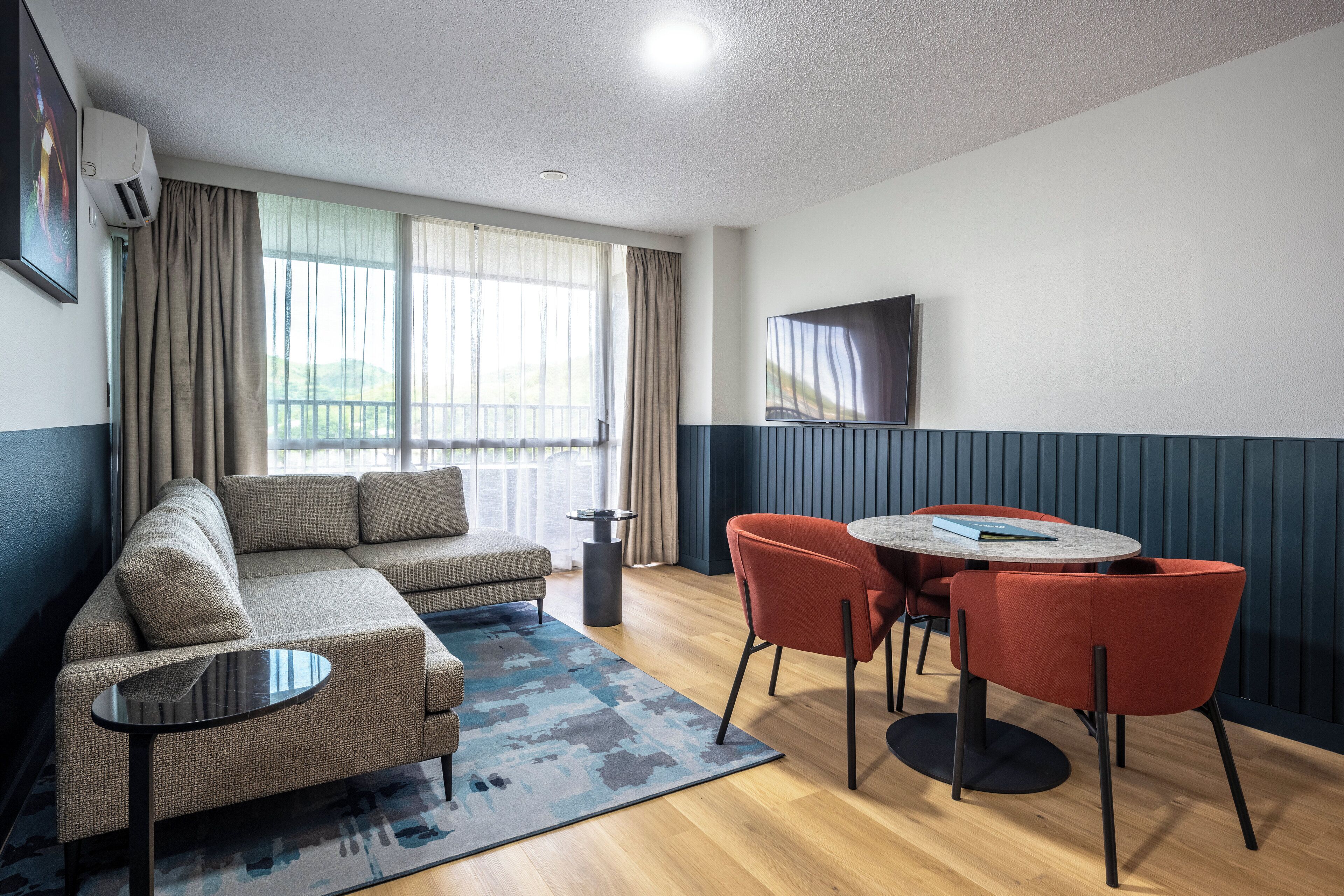 A modern living room with a sofa, a coffee table, and two chairs. There is a large window with curtains, a flat-screen TV mounted on the wall, and a ceiling light.