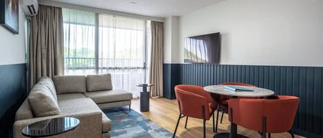A modern living room with a sofa, a coffee table, and two chairs. There is a large window with curtains, a flat-screen TV mounted on the wall, and a ceiling light.