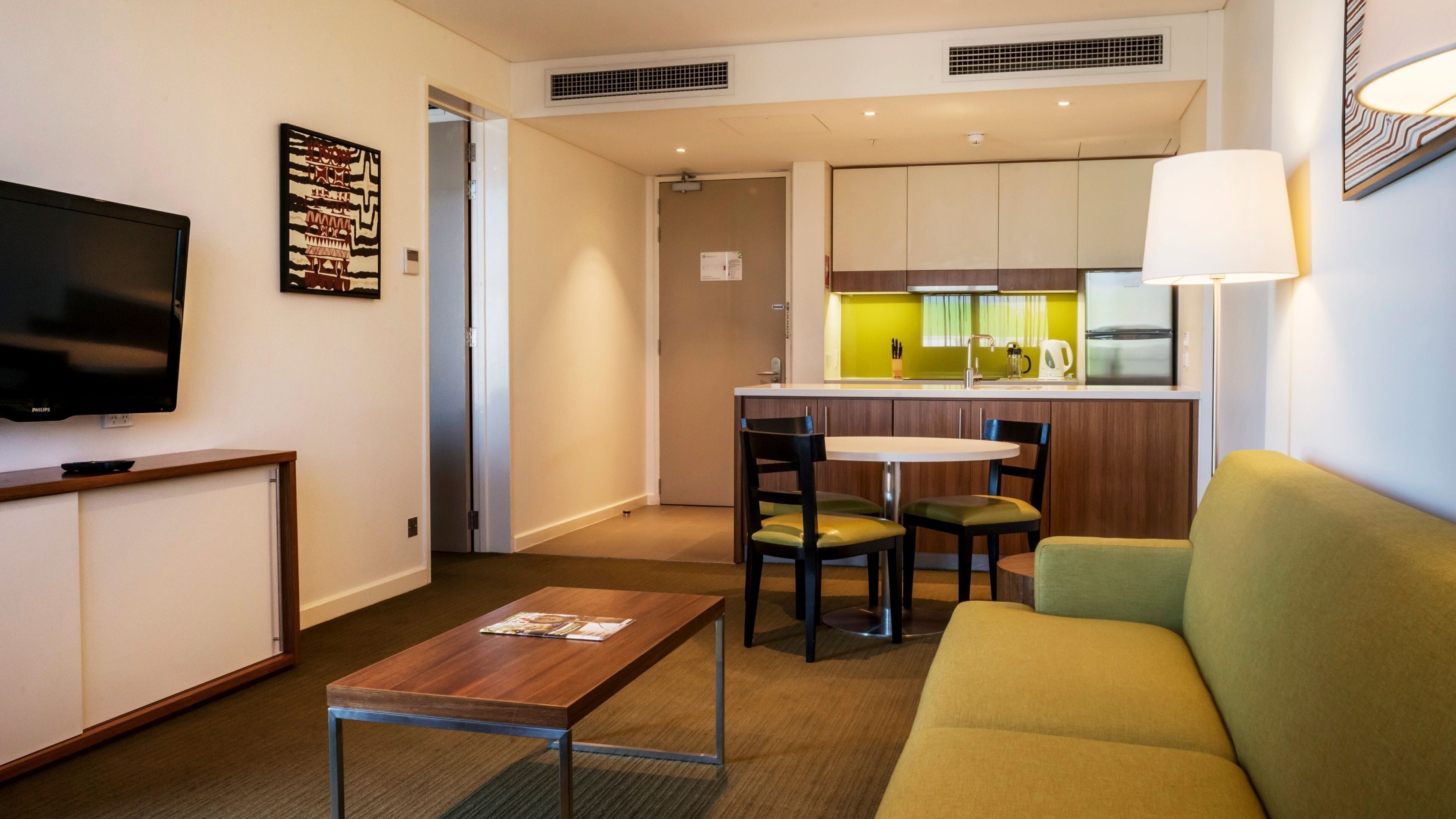 A hotel room with a TV, a wooden table, a green sofa, a small kitchen area, and a dining table with chairs.
