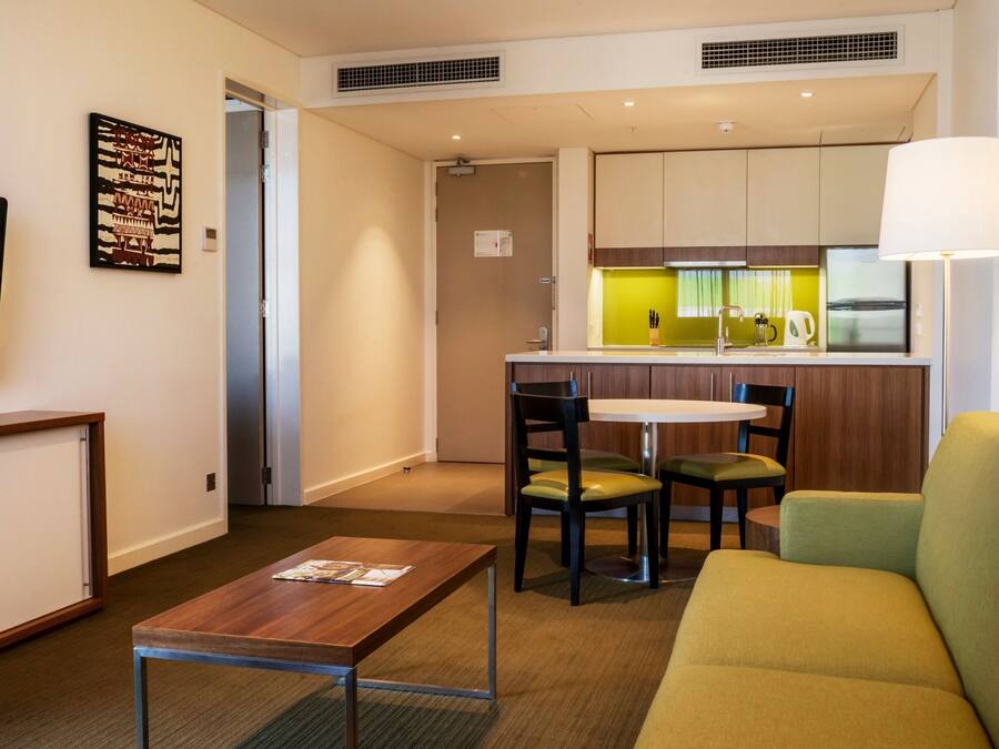 A hotel room with a TV, a wooden table, a green sofa, a small kitchen area, and a dining table with chairs.