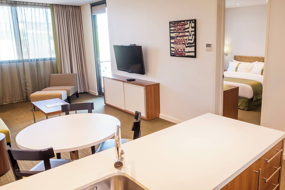 A hotel room with a round dining table, a sofa, a TV, and a bed with a headboard.
