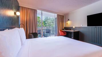 A hotel room with a large bed, a desk with a chair, a TV, and a balcony with a view of greenery.
