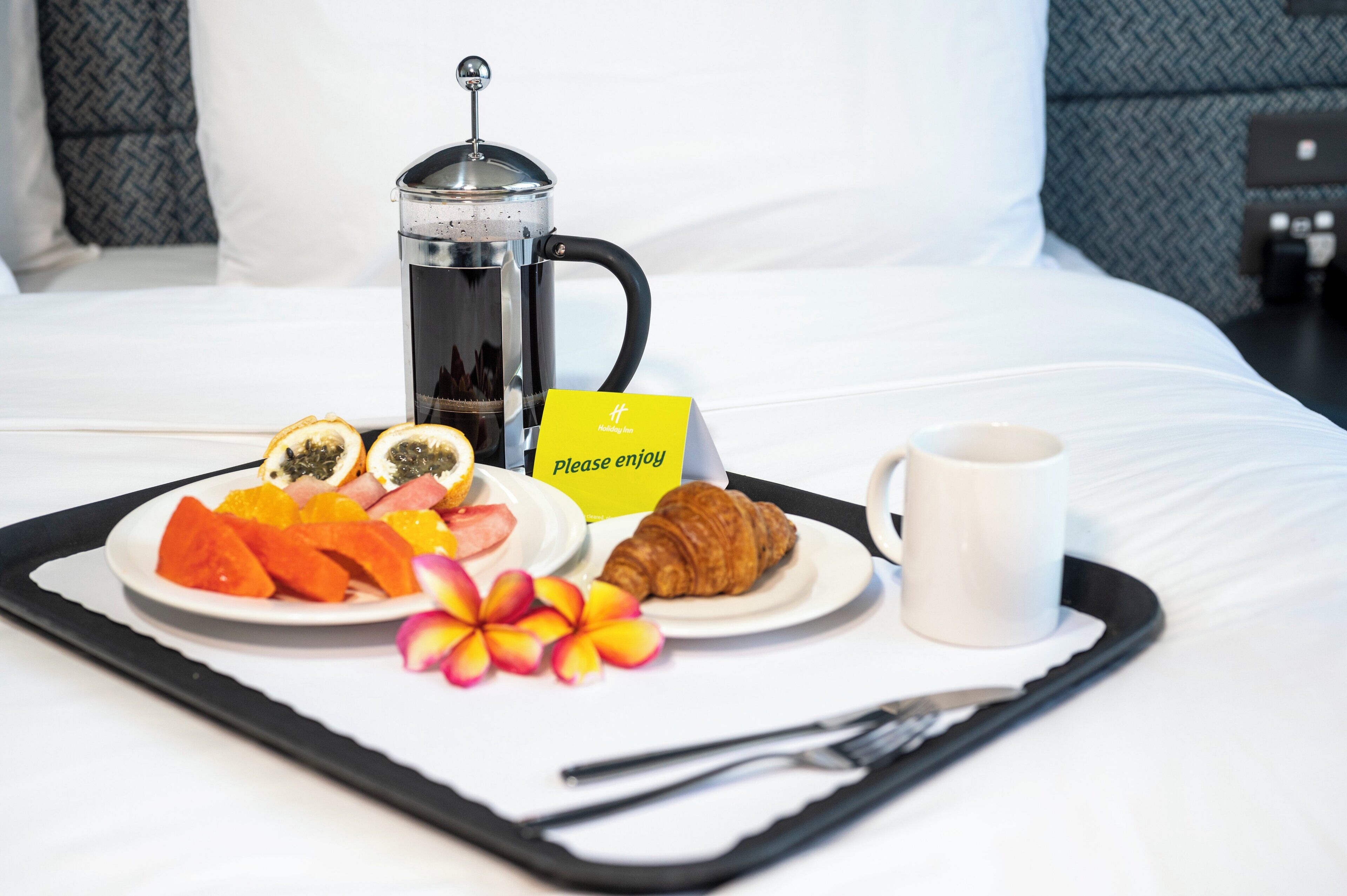 Un vassoio della colazione con una caffettiera, un croissant, frutta e un bigliettino su un letto.