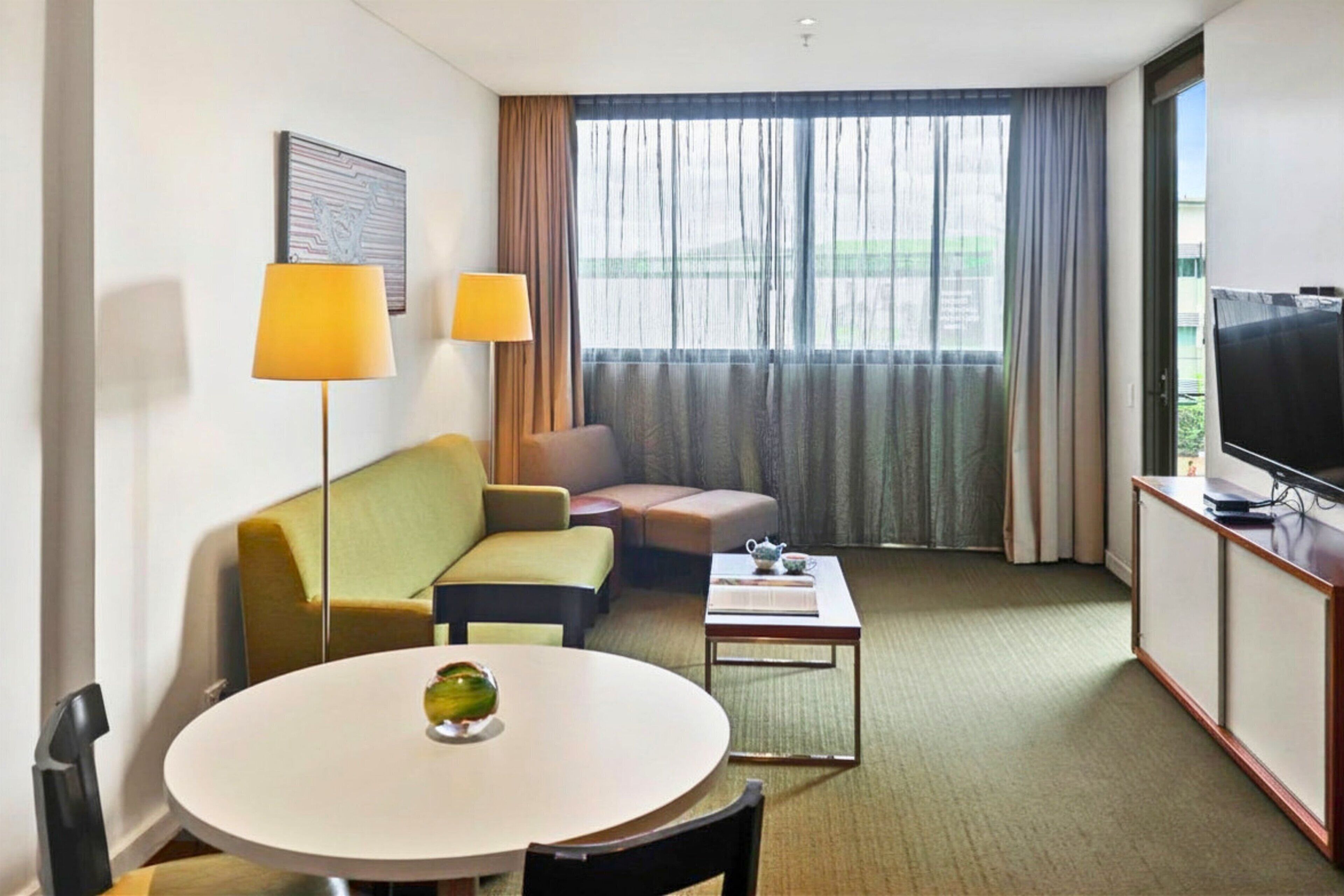 Habitación de hotel con una mesa de comedor redonda, un sofá, una televisión y una ventana con cortinas.